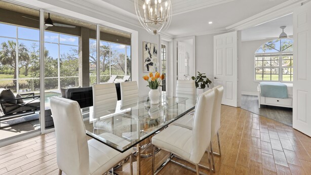 Um quarto espaçoso com mesa de jantar de vidro, cadeiras brancas, um lustre e amplas janelas que oferecem vista para o exterior.
