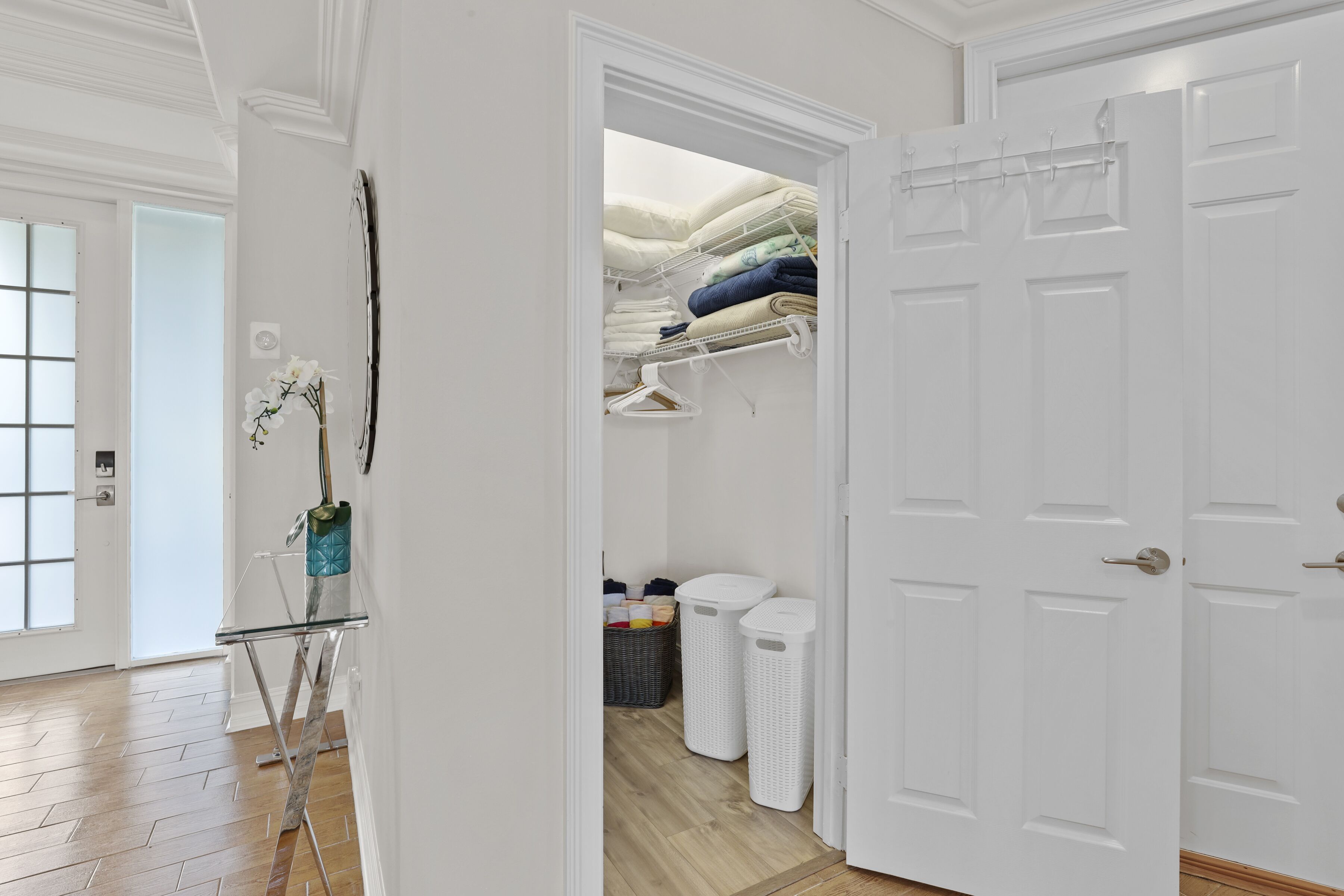 Un corridoio con pavimento in legno, un asse da stiro in vetro, un vaso con fiori e una porta bianca che conduce a una cabina armadio con scaffali e abiti appesi.