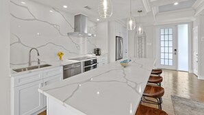 Uma cozinha moderna com armários brancos, uma ilha grande com banquetas, eletrodomésticos de aço inoxidável e iluminação pendente.