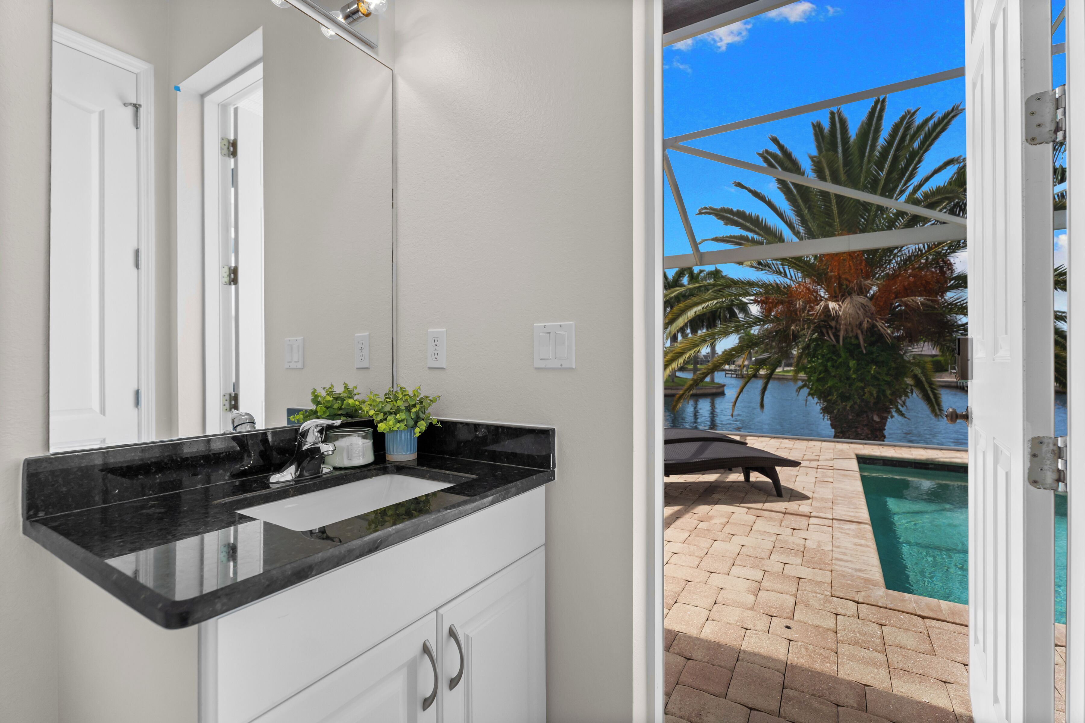 A bathroom with a black countertop, white cabinets, and a view of a pool and palm trees.