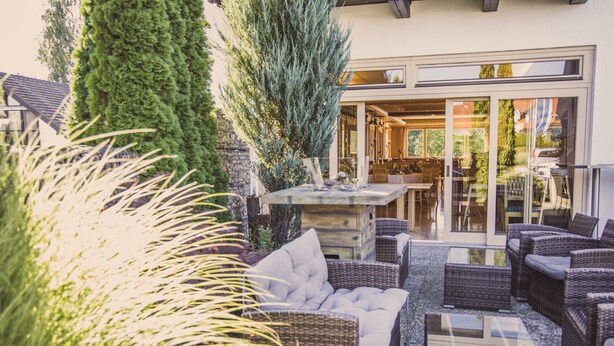 An outdoor seating area with wicker furniture and a stone table, surrounded by greenery and a building entrance. - Hotel & Gasthof Wagner (Aichach)