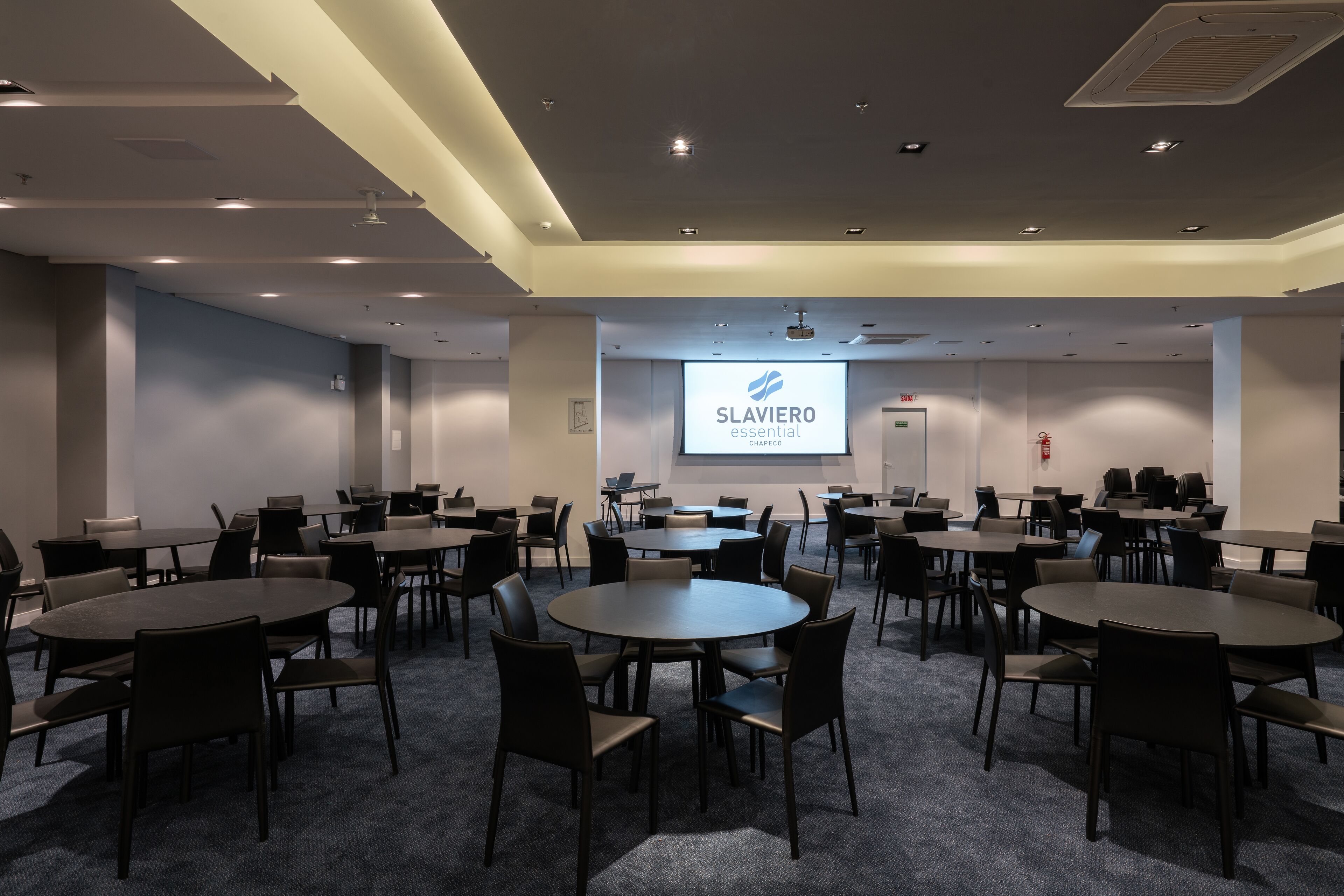 Un salón de conferencias con varias mesas y sillas, una pantalla de proyector mostrando 'SLAVIERO essential CHAPEGCO' y un extintor en la pared.