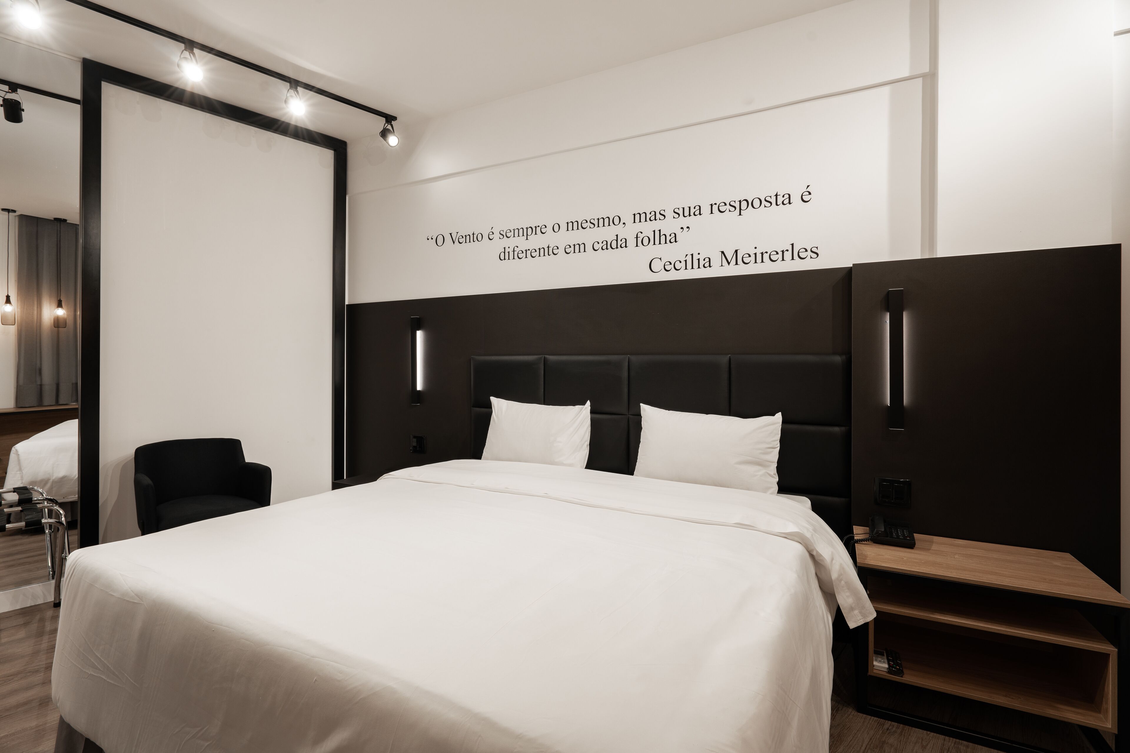A modern hotel room with a large bed, a black headboard, a bedside table with a lamp, and a quote on the wall.