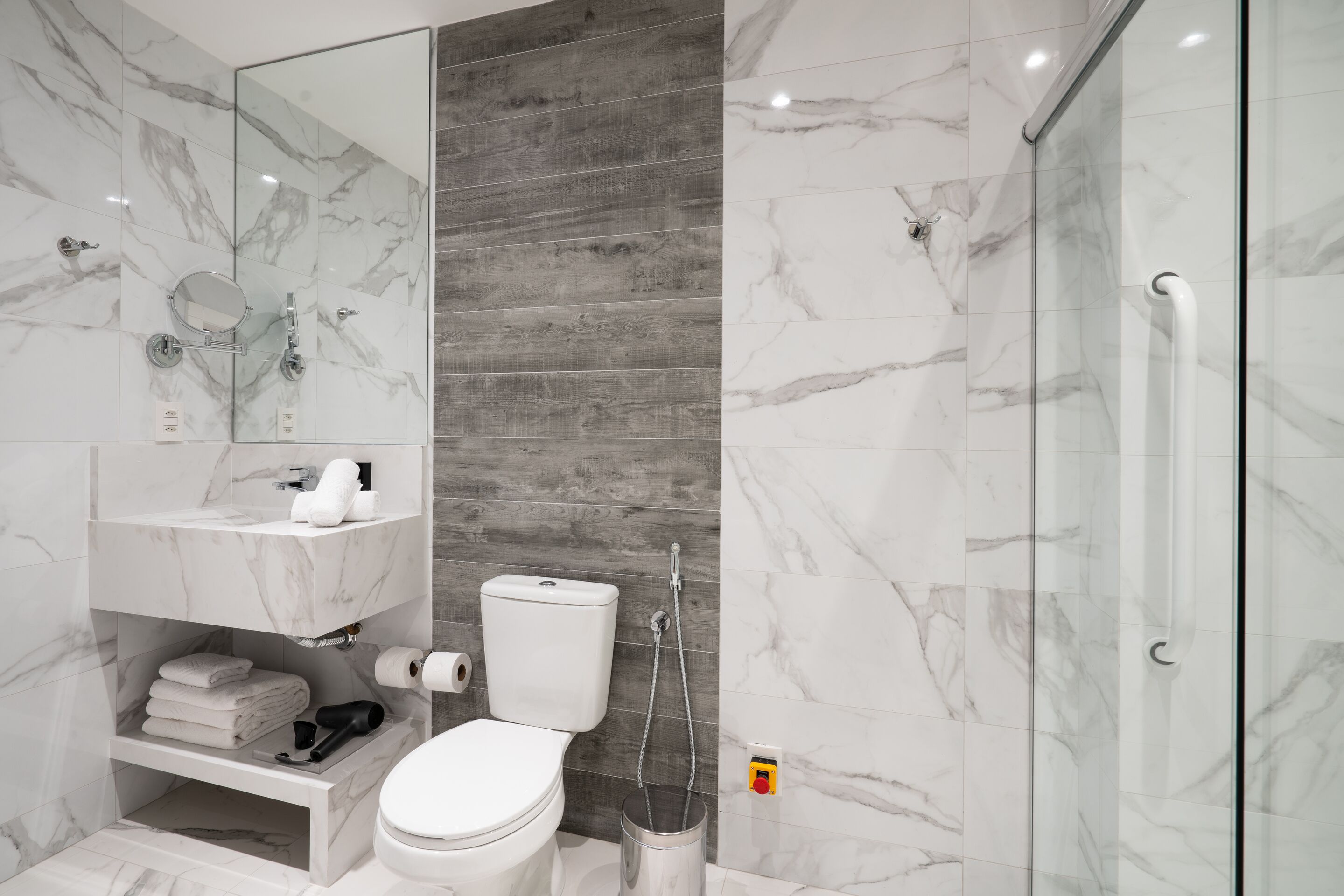 A modern bathroom with a white toilet, marble tiles, a wooden panel wall, a glass shower enclosure, and a white sink with a mirror above it.