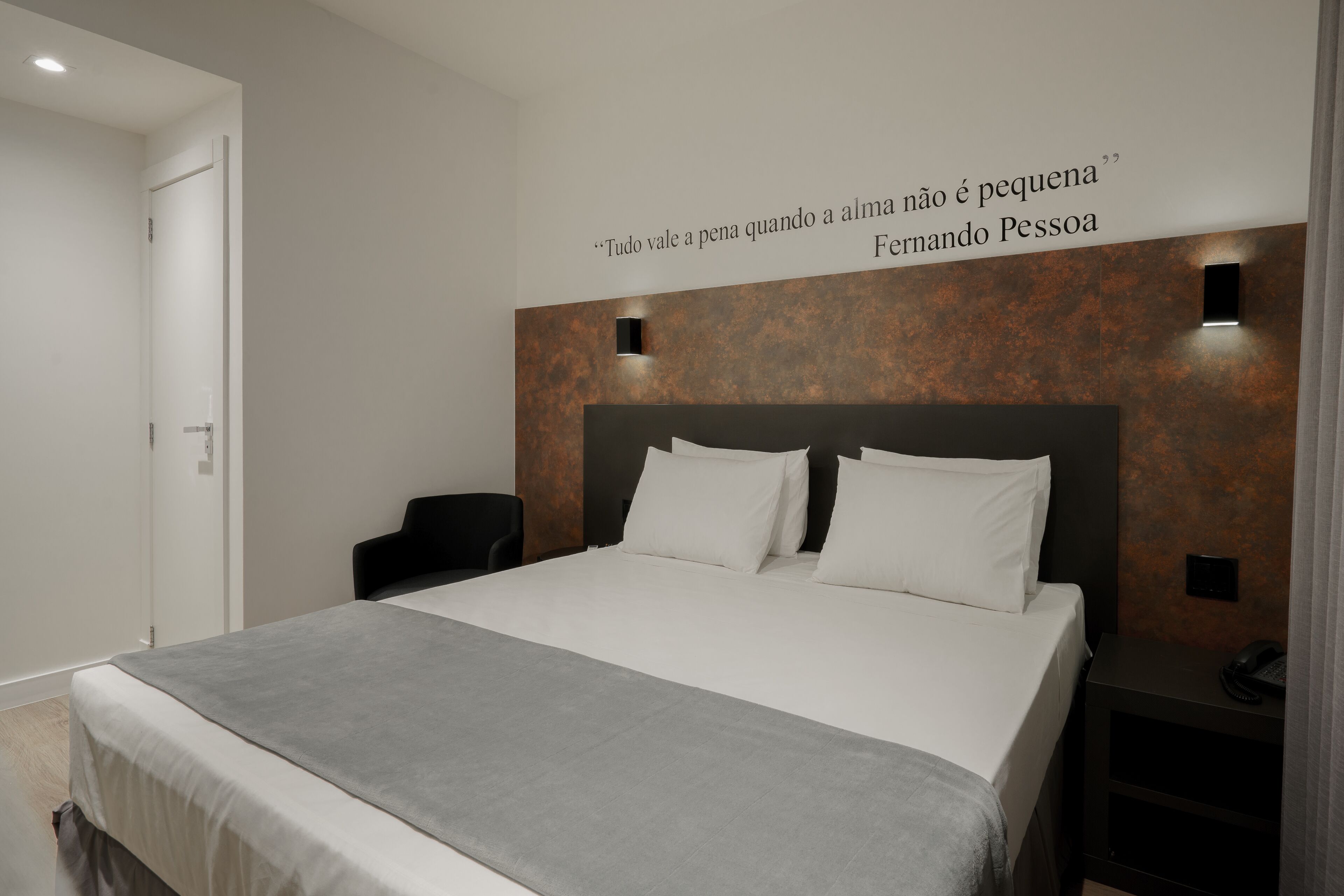 A modern hotel room with a large bed, a bedside table with a phone, and a quote on the wall.