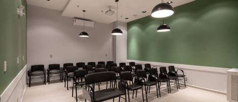 Un salón de conferencias con hileras de sillas negras, una pared verde grande y luces instaladas en el techo.