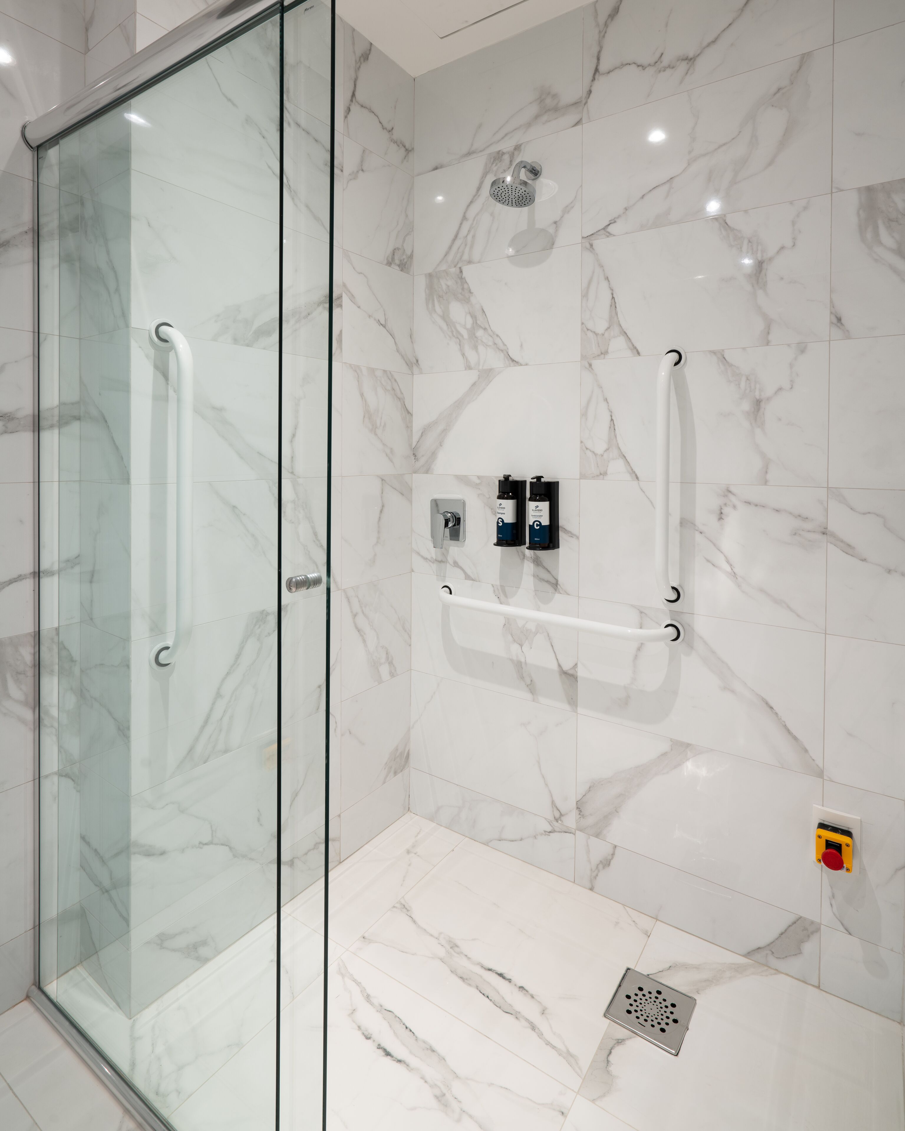 A modern bathroom with a glass shower enclosure, marble tiles, a handheld showerhead, and two soap dispensers.