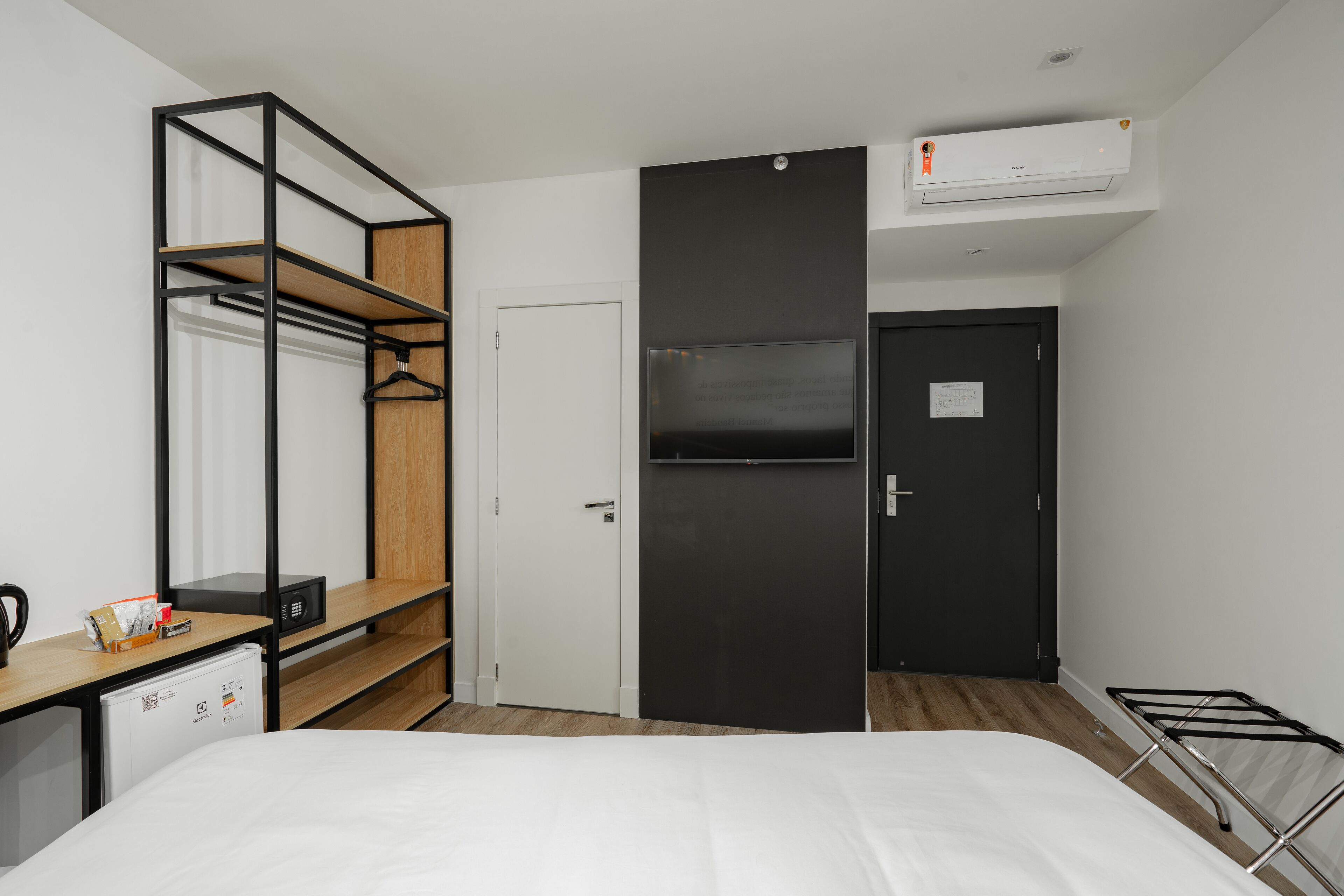 A compact hotel room with a bed, a wardrobe, a desk with a computer, and a small kitchenette.