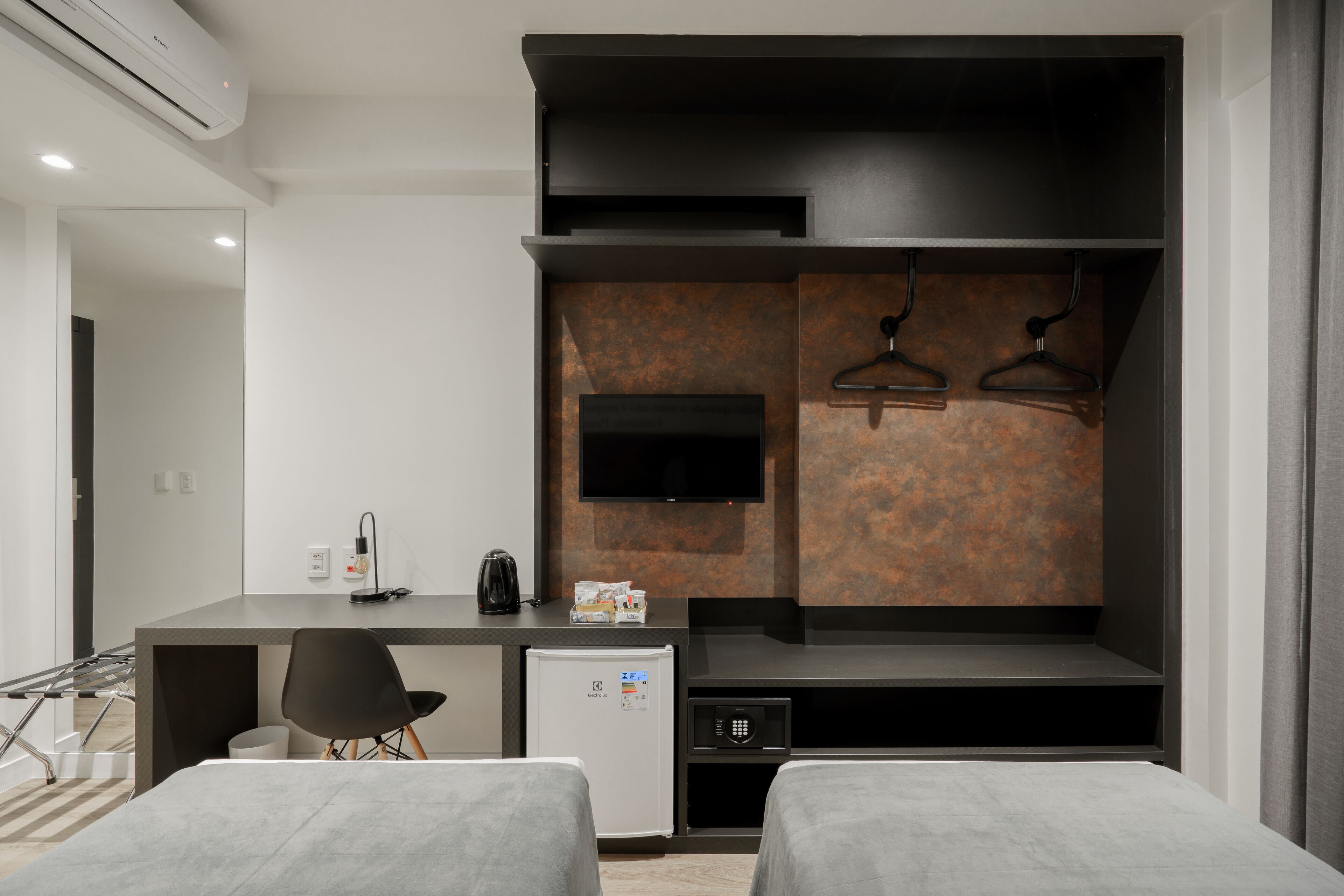 Habitación de hotel moderna con kitchenette, un televisor de pantalla plana montado en la pared, un refrigerador pequeño y una cama con ropa de cama gris.