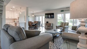 A spacious living area with a sofa, armchairs, a coffee table, and a dining table with chairs. There is a kitchen in the background with white cabinets and pendant lighting.