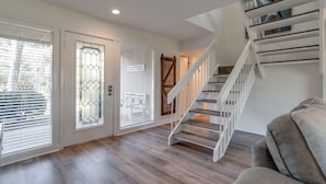 A spacious living room with a wooden floor, a staircase with white railings, and a door with a glass panel.