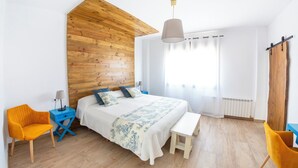 Un dormitorio con techo inclinado de madera, una cama con colcha blanca y azul estampada, una mesita de noche con lámpara y un banco.