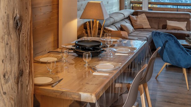 A wooden dining table with clear plastic covers, surrounded by chairs, in a room with exposed wooden beams and a large window.