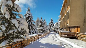 A snow-covered pathway flanked by wooden fences and snow-laden trees, leading to a modern building with balconies.