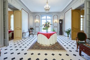 A room with a round table, a chandelier, a clock, and a large window. - Large family Suite garden view (Barbeville)