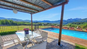 Zona de piscina con mesa y sillas de vidrio, una pérgola arriba y vistas a montañas y vegetación.