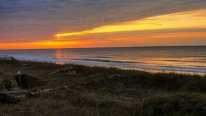 En solnedgång över en strand med en träpromenad och gräsklädda sanddyner.
