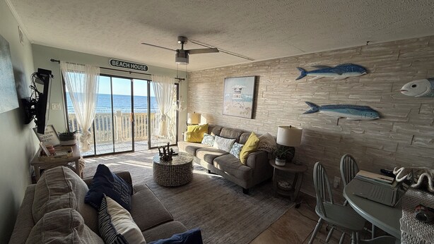 Ett vardagsrum med en soffa, fåtöljer, ett soffbord och ett matbord. Det finns en skylt med texten ”STRANDHUS” och en utsikt över havet genom stora fönster.