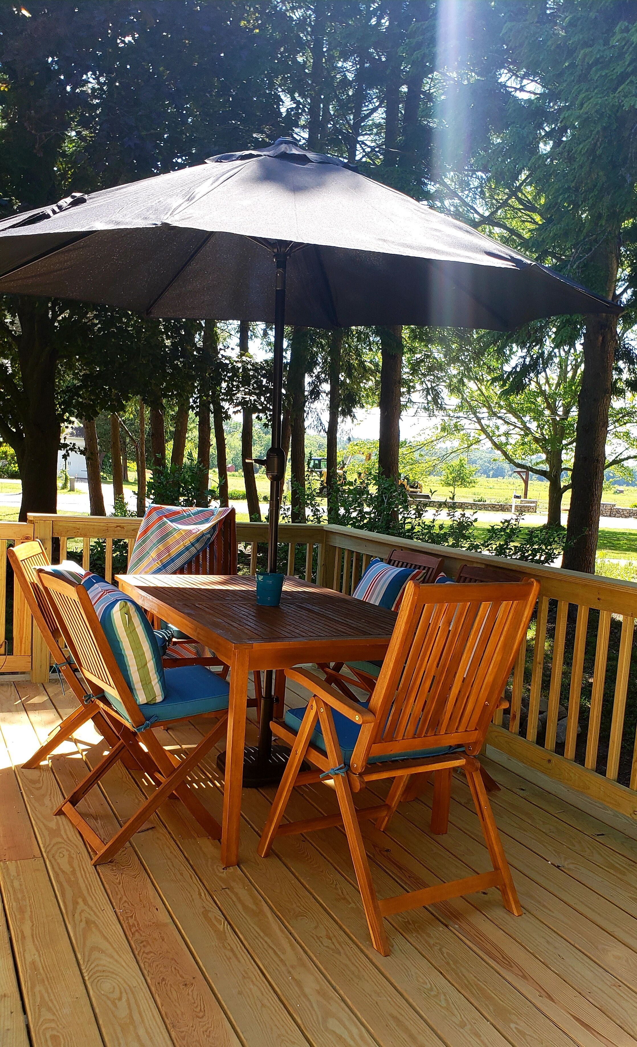 Una terraza de madera al aire libre con una mesa, sillas y un gran paraguas, con vistas a un paisaje verde.