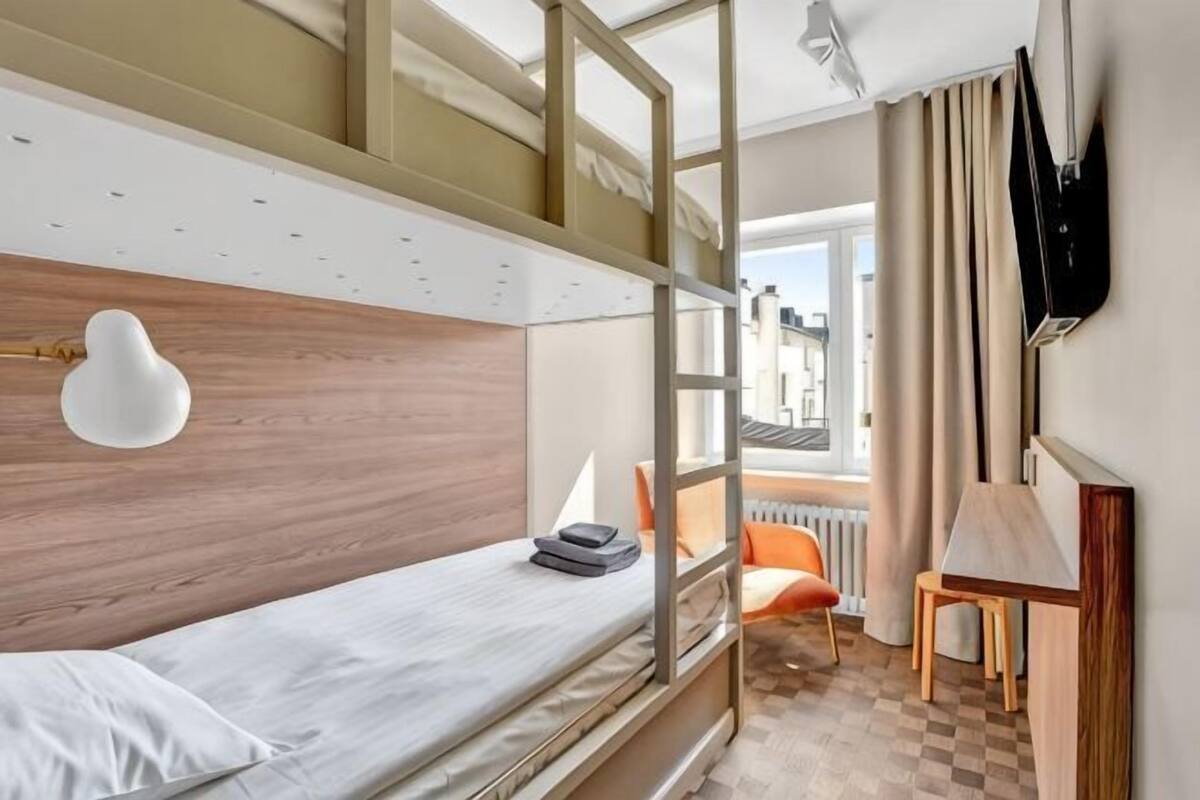 Une chambre d’hôtel moderne équipée d’un lit superposé, d’un bureau avec une chaise, d’un téléviseur fixé au mur et d’une fenêtre avec des rideaux.
