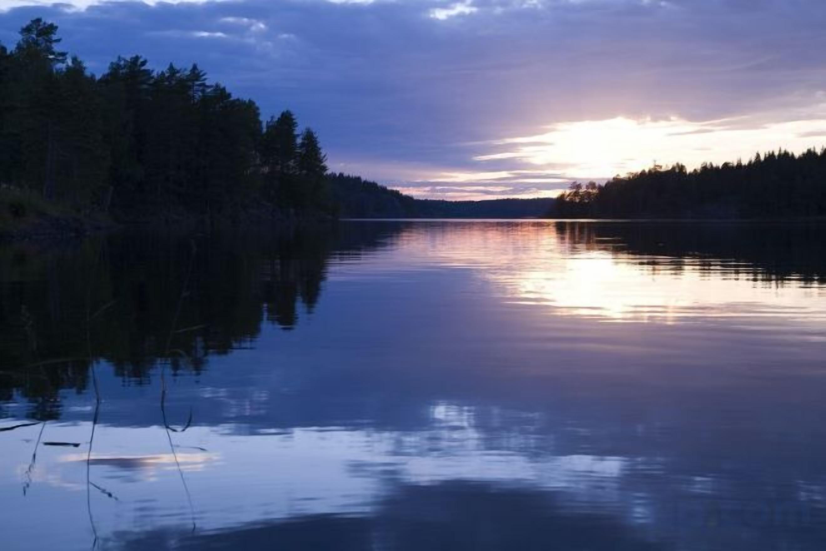 A calm lake with a forested shoreline and a sunset in the background.