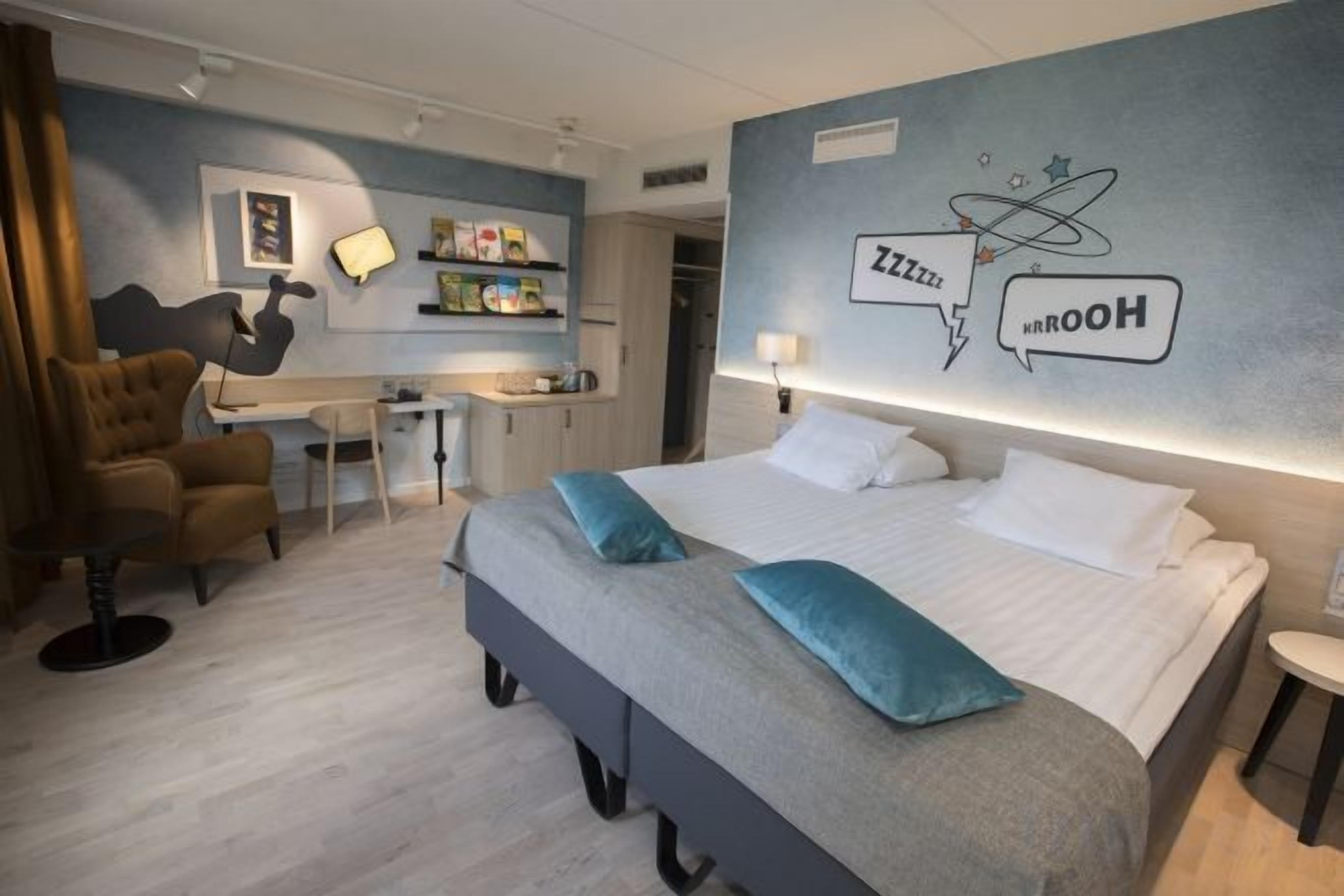 Una habitación de hotel con cama, escritorio, silla y pared con un diseño gráfico.
