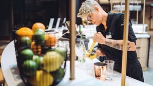 Un barman prépare un cocktail dans un bar, avec une variété de fruits et de bouteilles en arrière-plan.