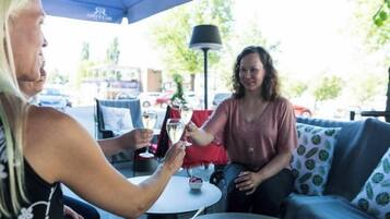 Two people seated at an outdoor table with a drink, under a blue umbrella, with a street and parked cars in the background.