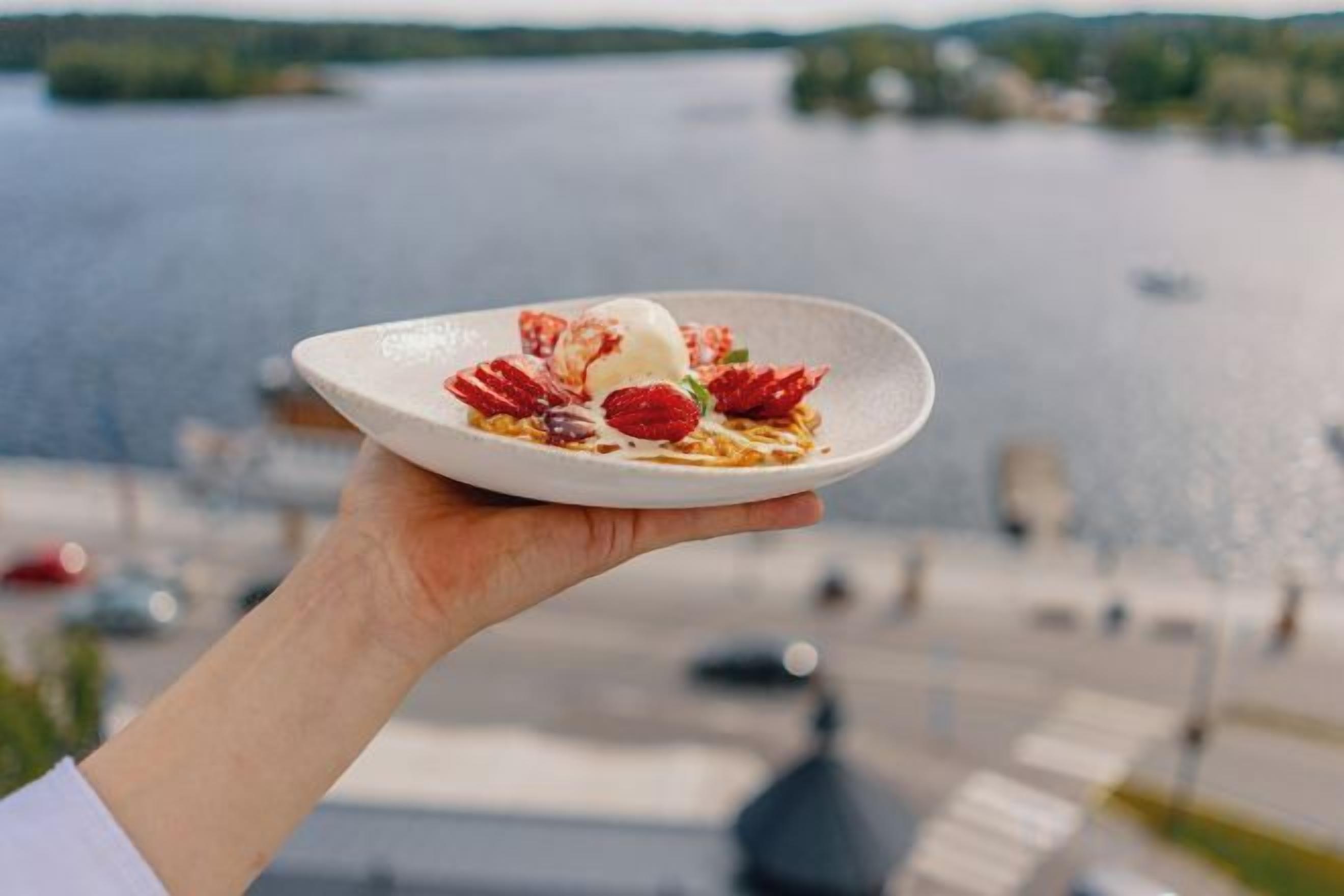 A hand holding a plate with a dessert against a backdrop of a body of water and a distant shoreline.