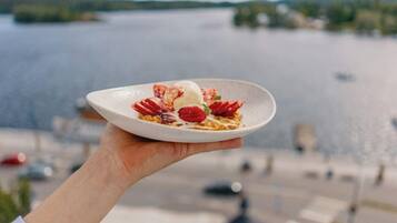 A hand holding a plate with a dessert against a backdrop of a body of water and a distant shoreline.