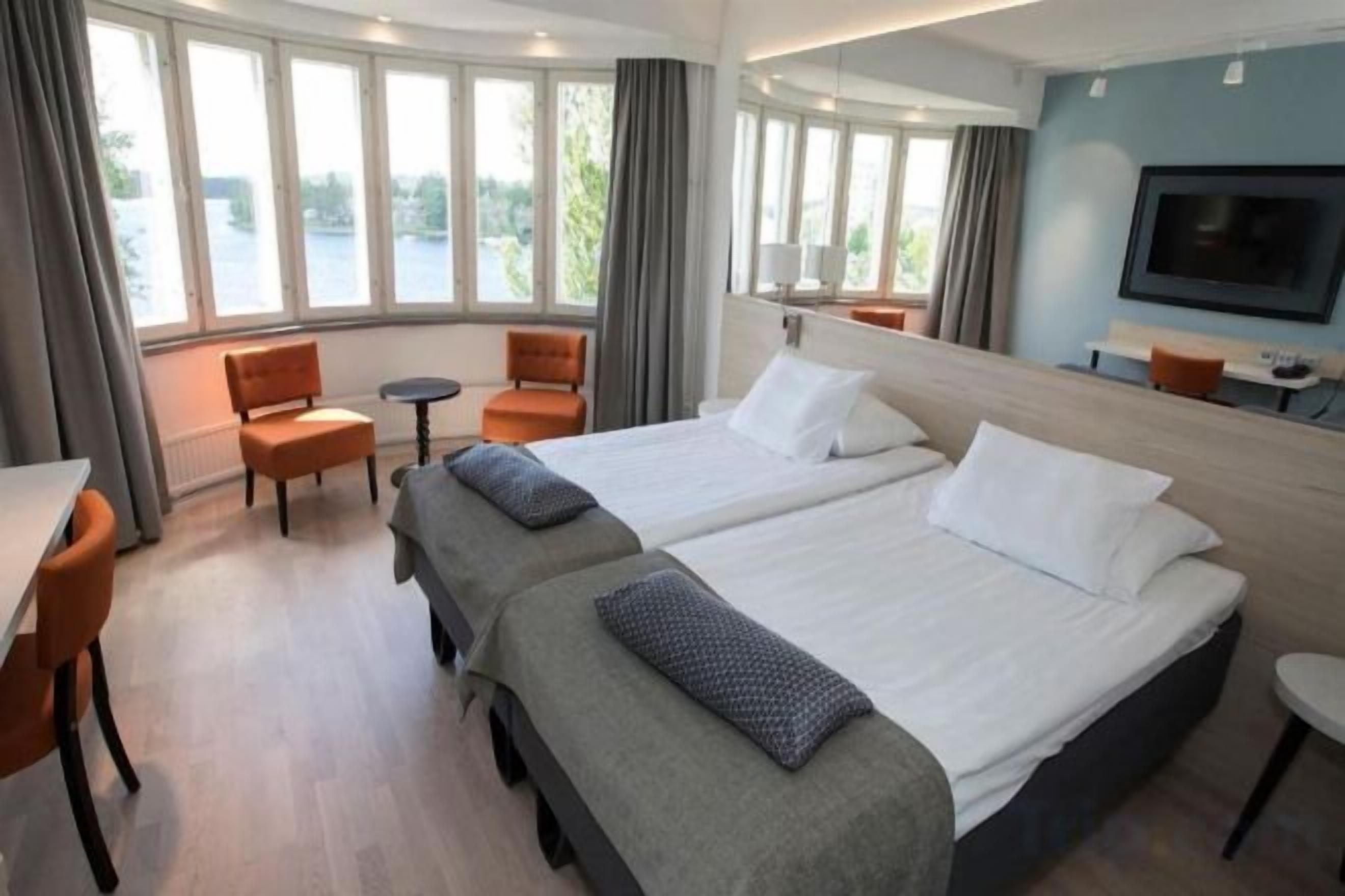 A hotel room with two beds, a desk, chairs, a TV, and large windows with a view.