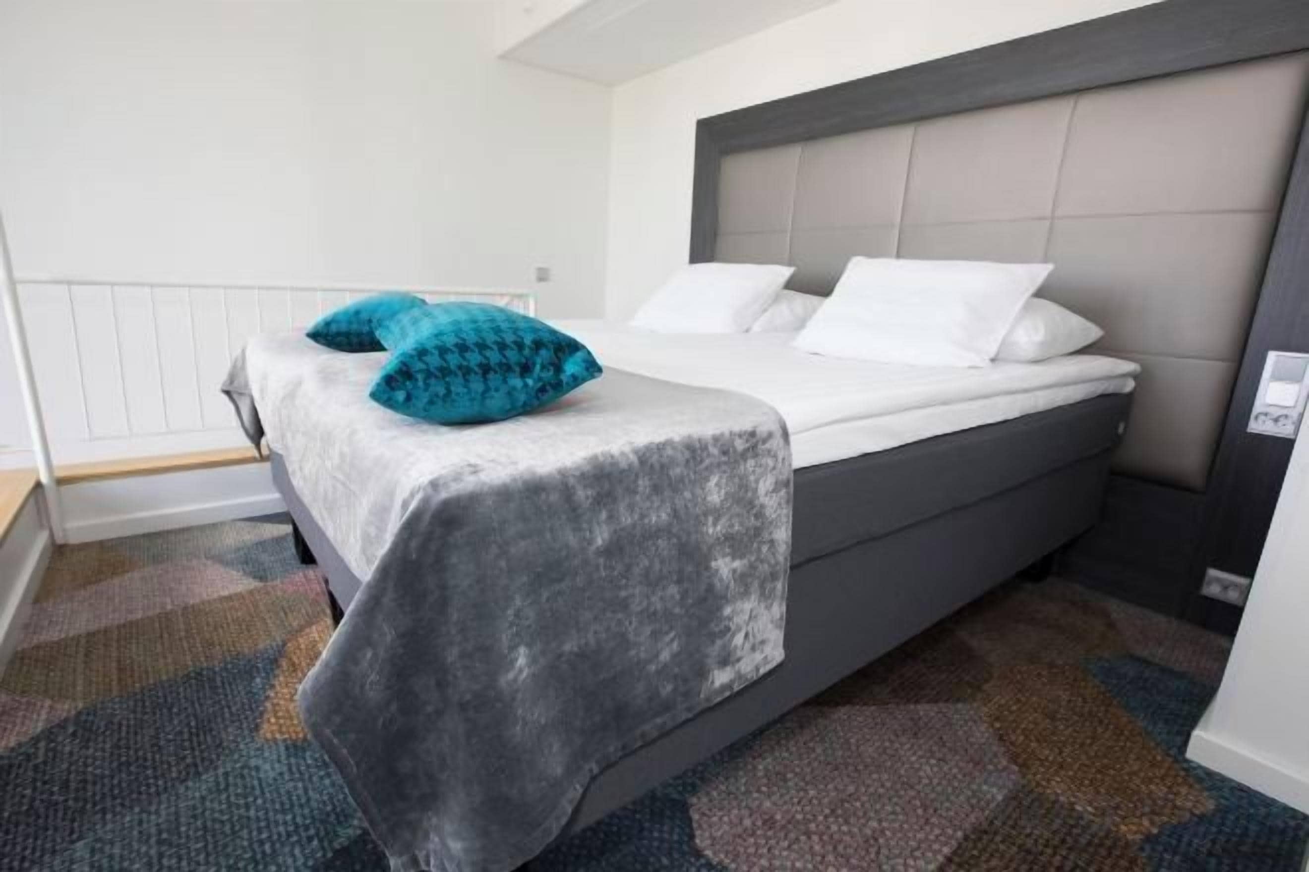 Ein Bett mit grauem Kopfteil, weißer Bettwäsche und einem gemusterten Teppich.