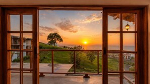 A view through a wooden-framed window with a metal railing, overlooking a sunset over a coastal landscape with a tree and buildings. - Family_friendly beach house  (Jama)