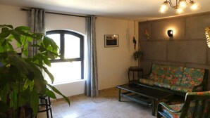 A living room with a sofa, a coffee table, a potted plant, a window with curtains, and a wall-mounted light fixture.