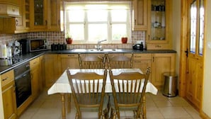 A kitchen with wooden cabinets, a dining table with chairs, a window with curtains, and a trash can. - Scenic Estuary Way 3-bed Home in Labasheeda (Labasheeda)