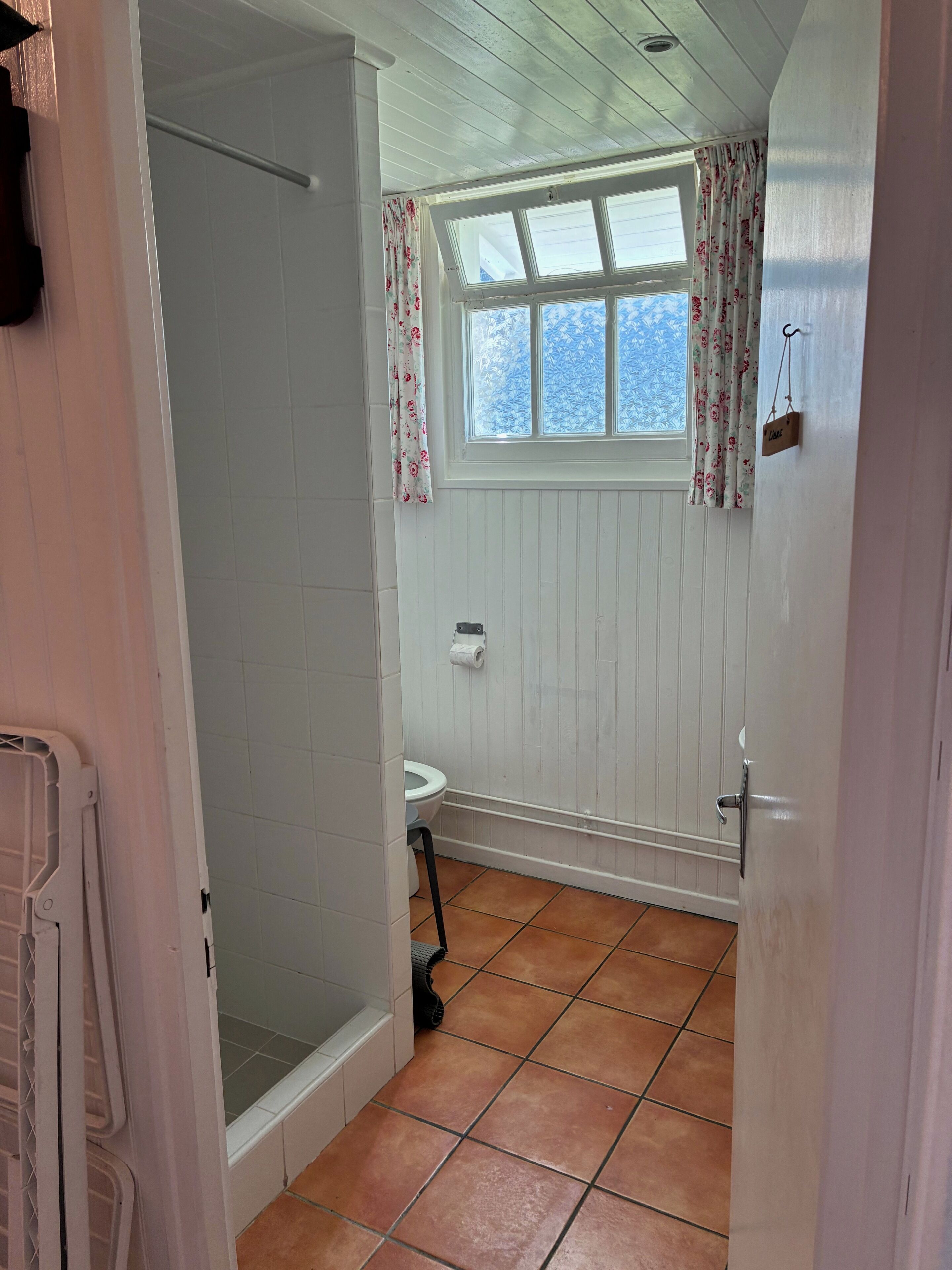 A bathroom with a shower, toilet, and window with curtains.