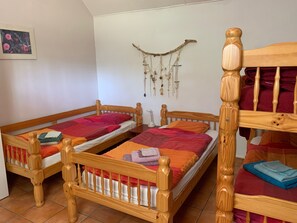 Une pièce avec deux lits en bois, chacun doté d’une tête de lit et d’un pied de lit. Un panneau mural en macramé et un tableau encadré sont accrochés au mur, au-dessus des lits.