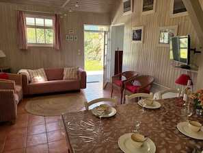 Un salon chaleureux avec un canapé, des fauteuils en osier, une table à manger dressée pour deux personnes et un téléviseur fixé au mur.