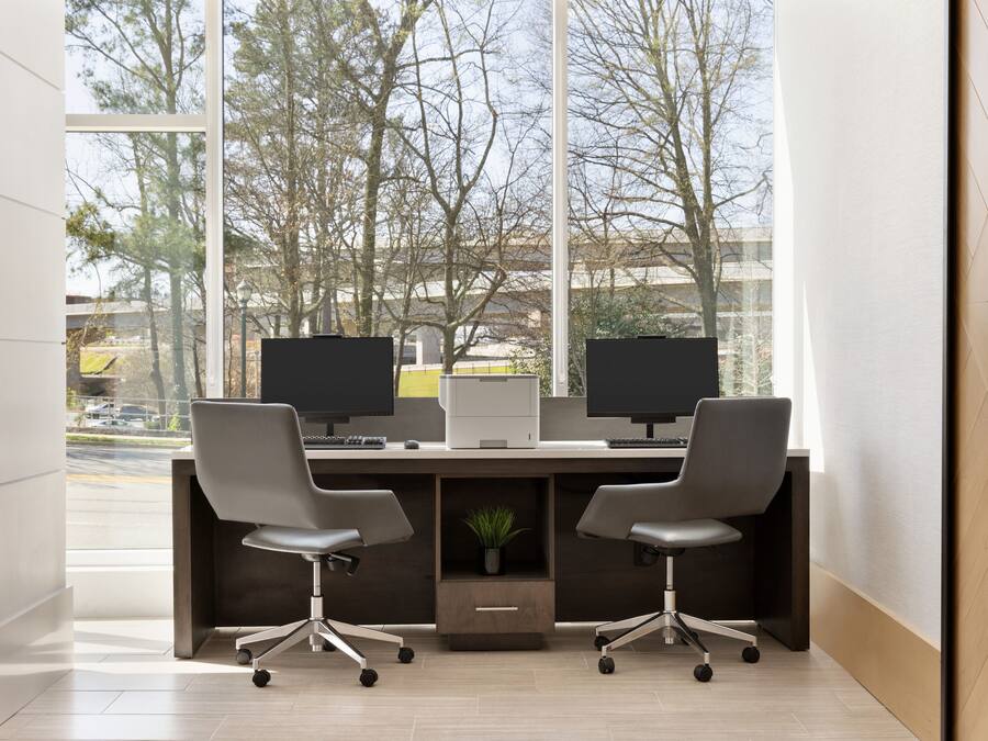 Een modern kantoor met twee bureaus, elk met een beeldscherm, toetsenbord en stoel. Op het bureau staat een kleine plant, en de kamer heeft grote ramen met uitzicht op bomen en gebouwen.