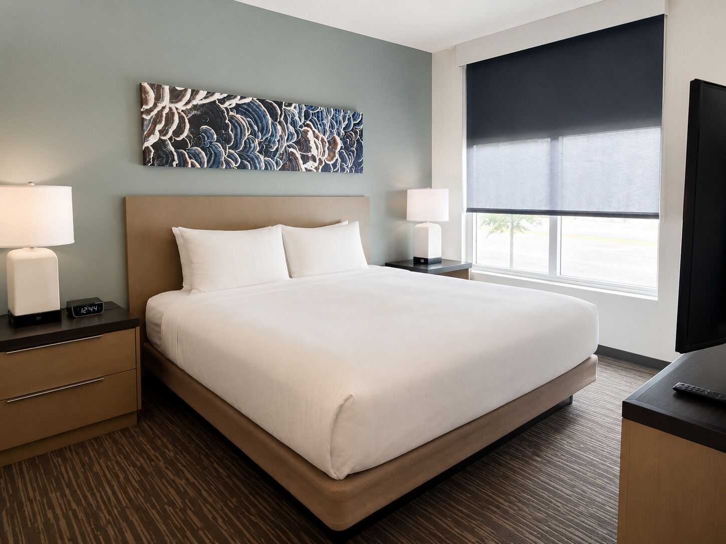 A hotel room with a large bed, two bedside tables with lamps, a wall art piece, and a window with a view.