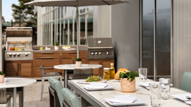 An outdoor dining area with a table set for two, a grill, and a view of the city. - Hyatt House Atlanta / Perimeter Center (Atlanta)