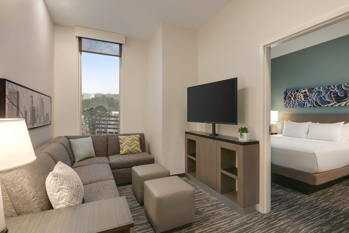 A hotel room with a large window, a flat-screen TV, a bed, a sofa, and a small table.