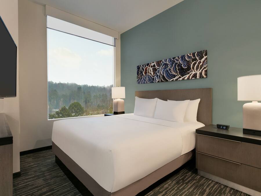 Quarto de hotel com uma cama grande, mesa de cabeceira com luminária, relógio de parede e vista para o exterior.