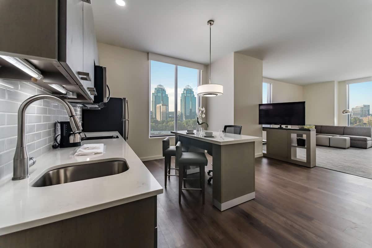 A modern kitchen with a large island, a dining area with bar stools, a flat-screen TV, and a city view through large windows.