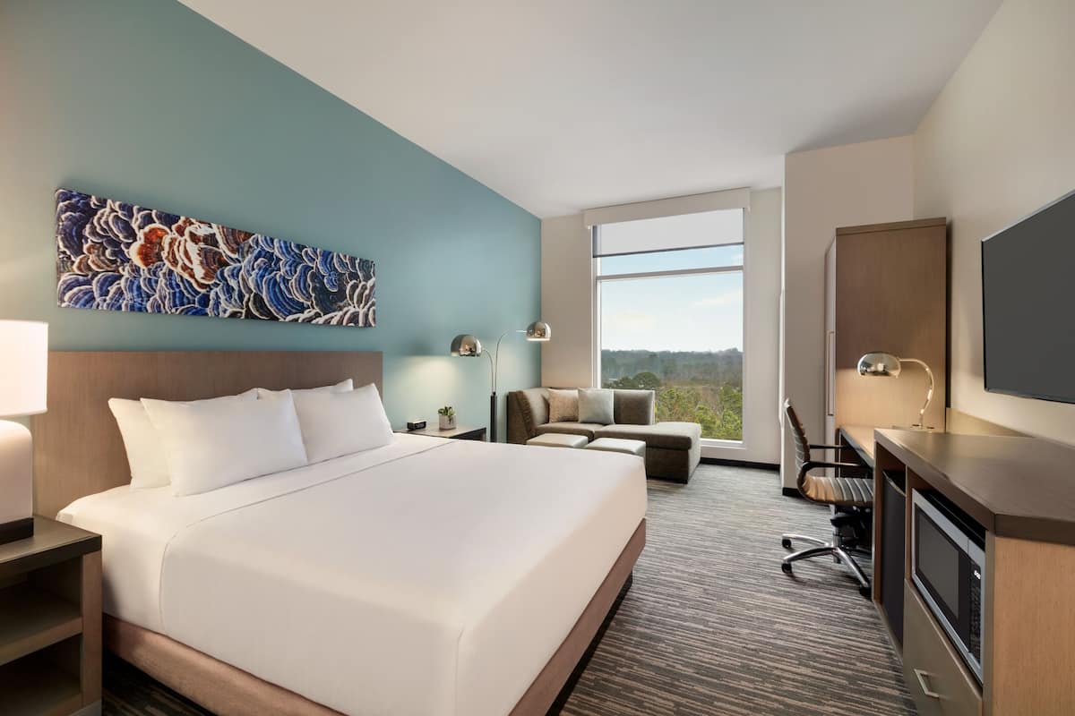 A hotel room with a large bed, a desk with a computer, a sofa, and a TV. There is a window with a view of the outdoors.