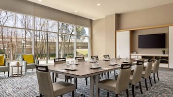 A conference room with a large table, chairs, a TV screen, and large windows offering a view of trees and buildings.