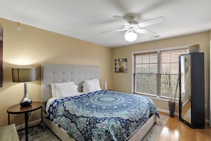 A bedroom with a bed, a ceiling fan, a nightstand with a lamp, a window with blinds, and a mirror.