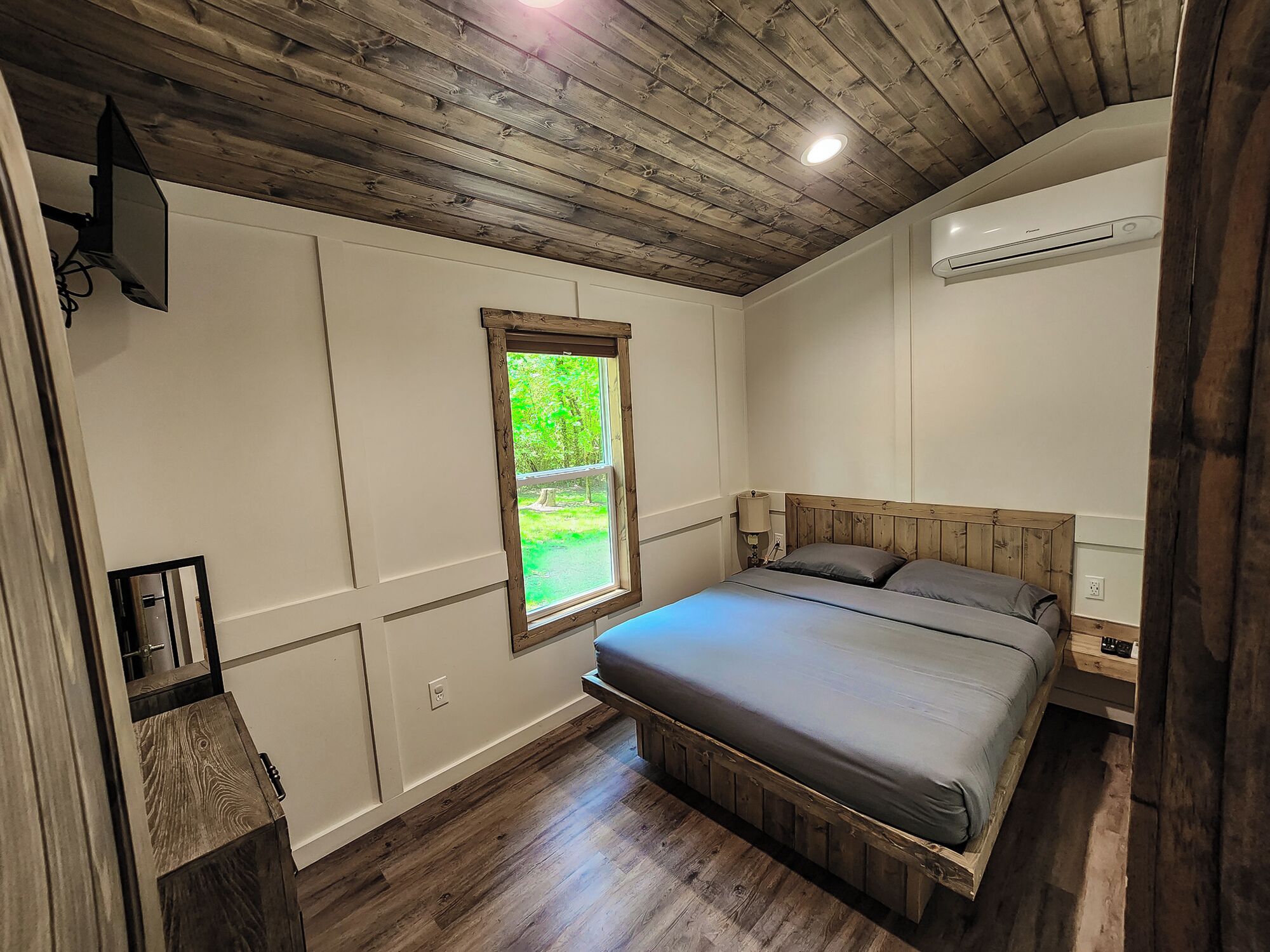 Une chambre avec un plafond en bois, une fenêtre donnant sur la verdure, un lit avec une tête de lit en bois et un appareil de climatisation.