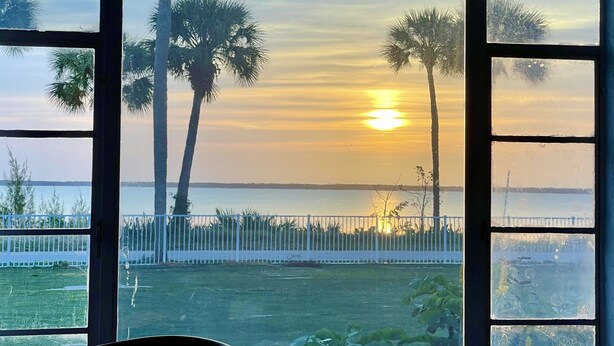 A view through a window with a white fence and palm trees, overlooking a body of water at sunset. - 3 Bedroom 3 Bath  a waterfront 3 Acre Home close to Downtown and Beaches. (Fort Pierce)