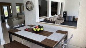 A dining area with a table set for four, a bowl of fruit, and a view of a living room with a sofa and a painting. - 3 Bedroom 3 Bath  a waterfront 3 Acre Home close to Downtown and Beaches. (Fort Pierce)